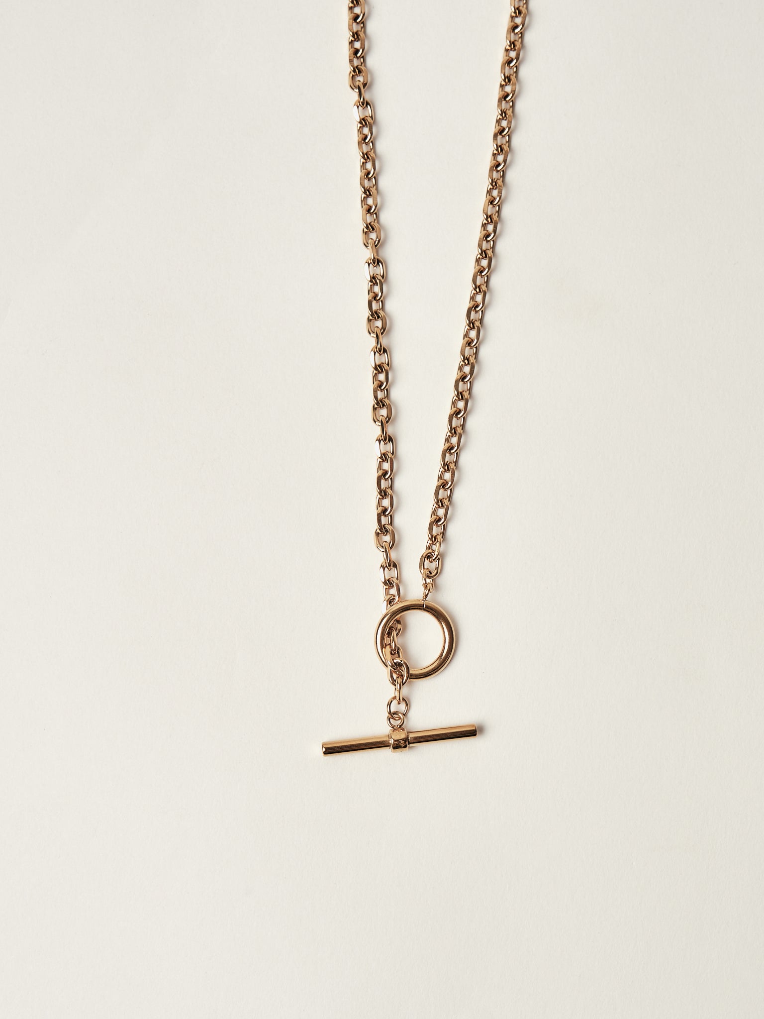 Gold necklace with a round pendant and a bar-shaped charm on a light beige background