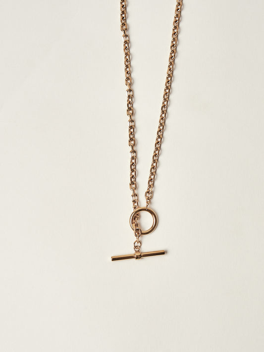 Gold necklace with a round pendant and a bar-shaped charm on a light beige background