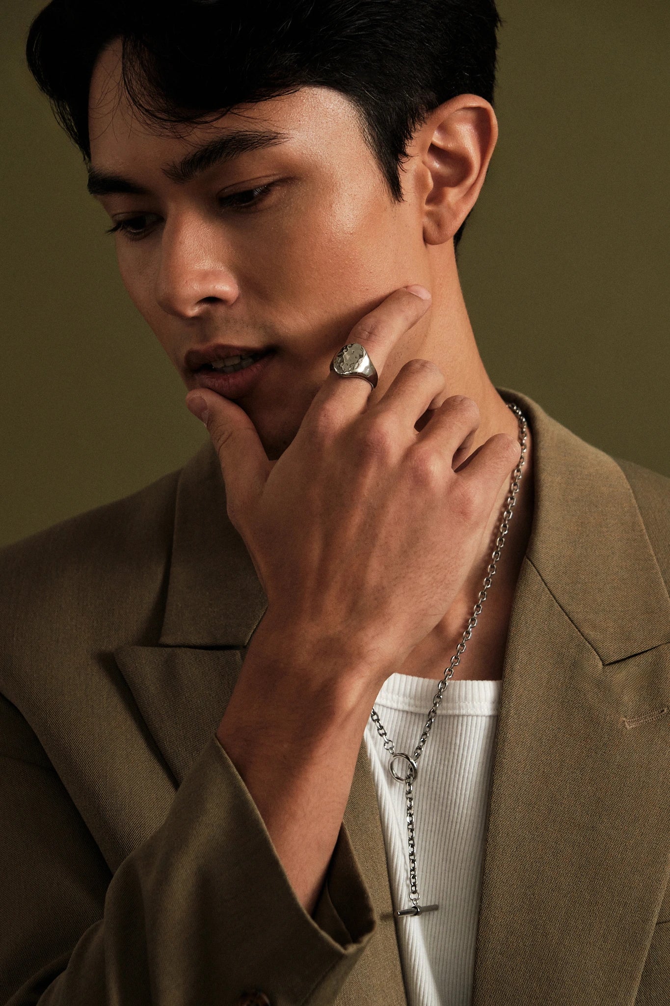 Man wearing a brown jacket and white shirt with a necklace, against a green background