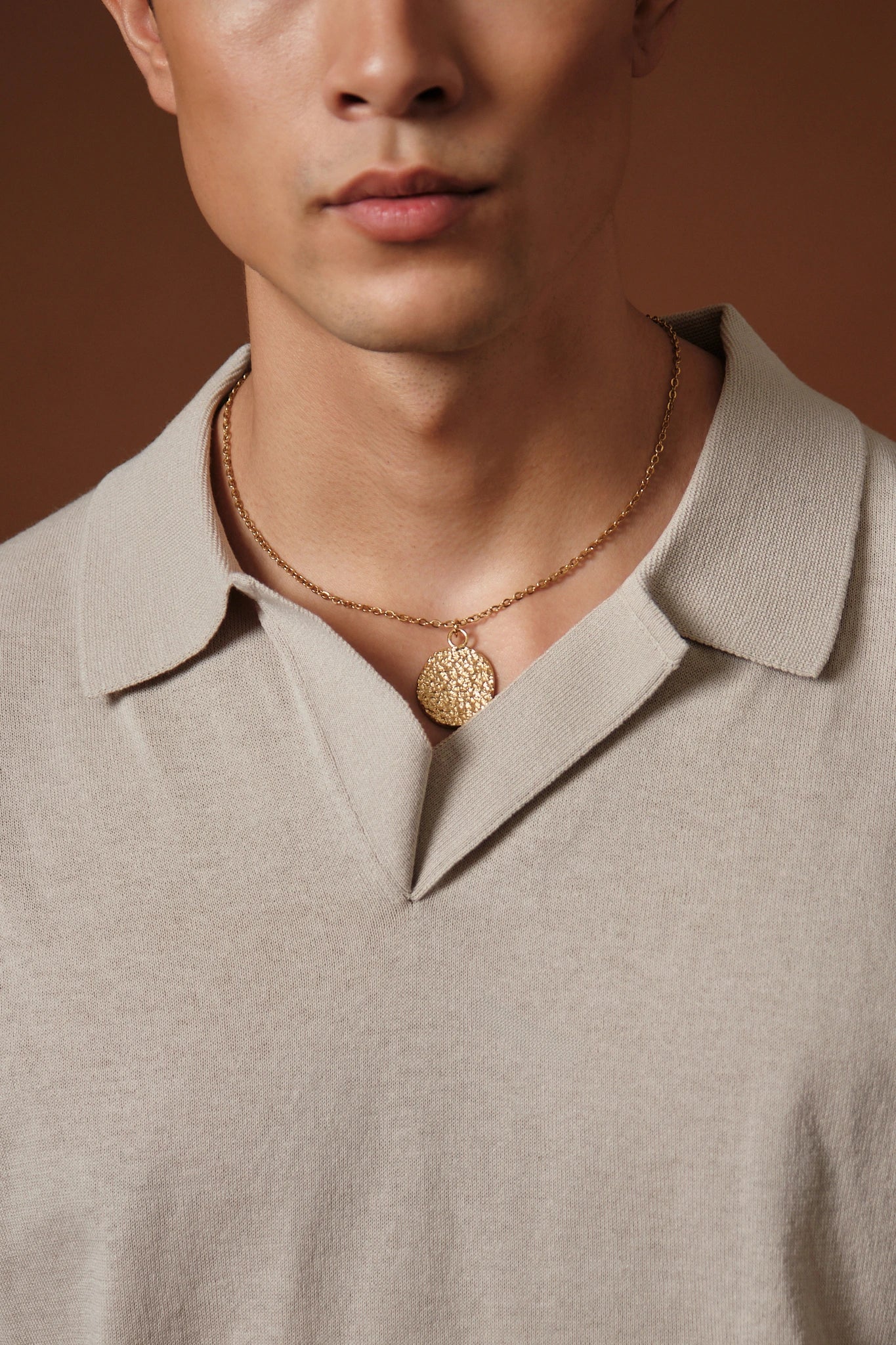 Man wearing a beige shirt with a gold necklace featuring a round pendant against a brown background