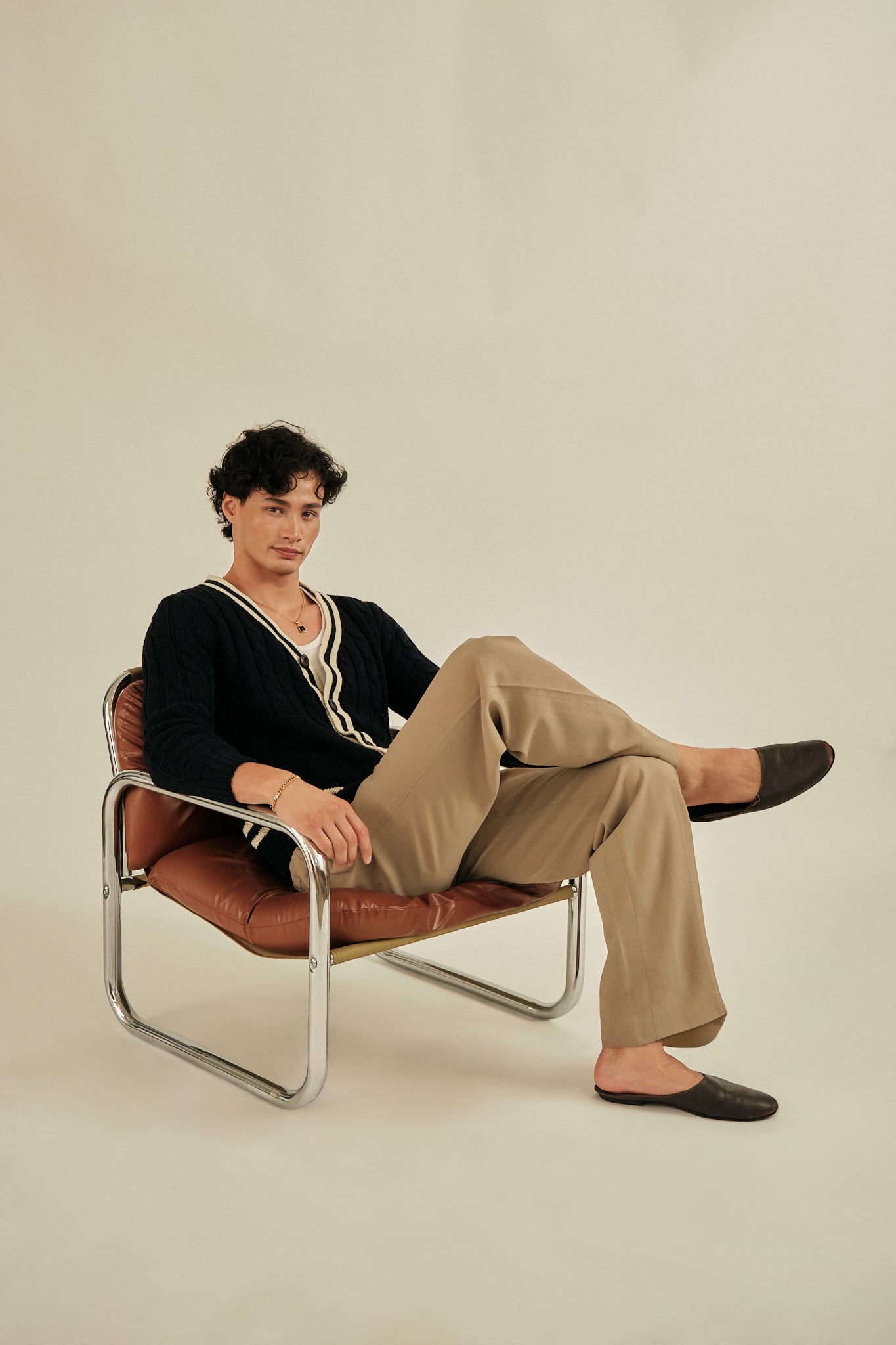 Person sitting on a chair wearing a black sweater and beige pants against a plain background