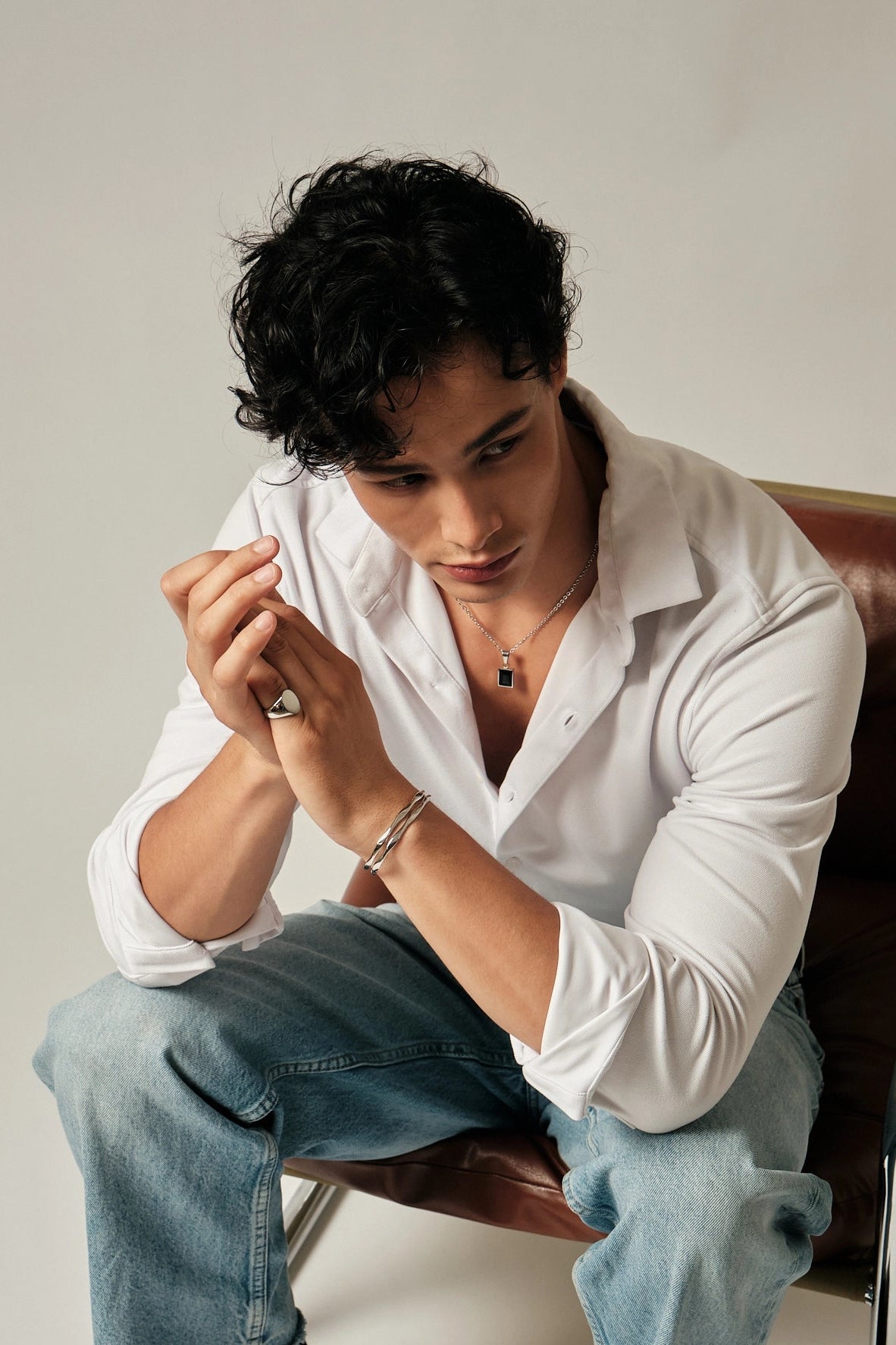 Man sitting on a chair wearing a white shirt and blue jeans, looking at his hands.