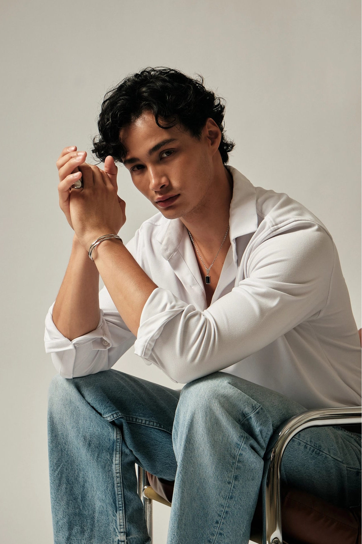 Man sitting on a chair wearing a white shirt and blue jeans, holding his hand to his face.