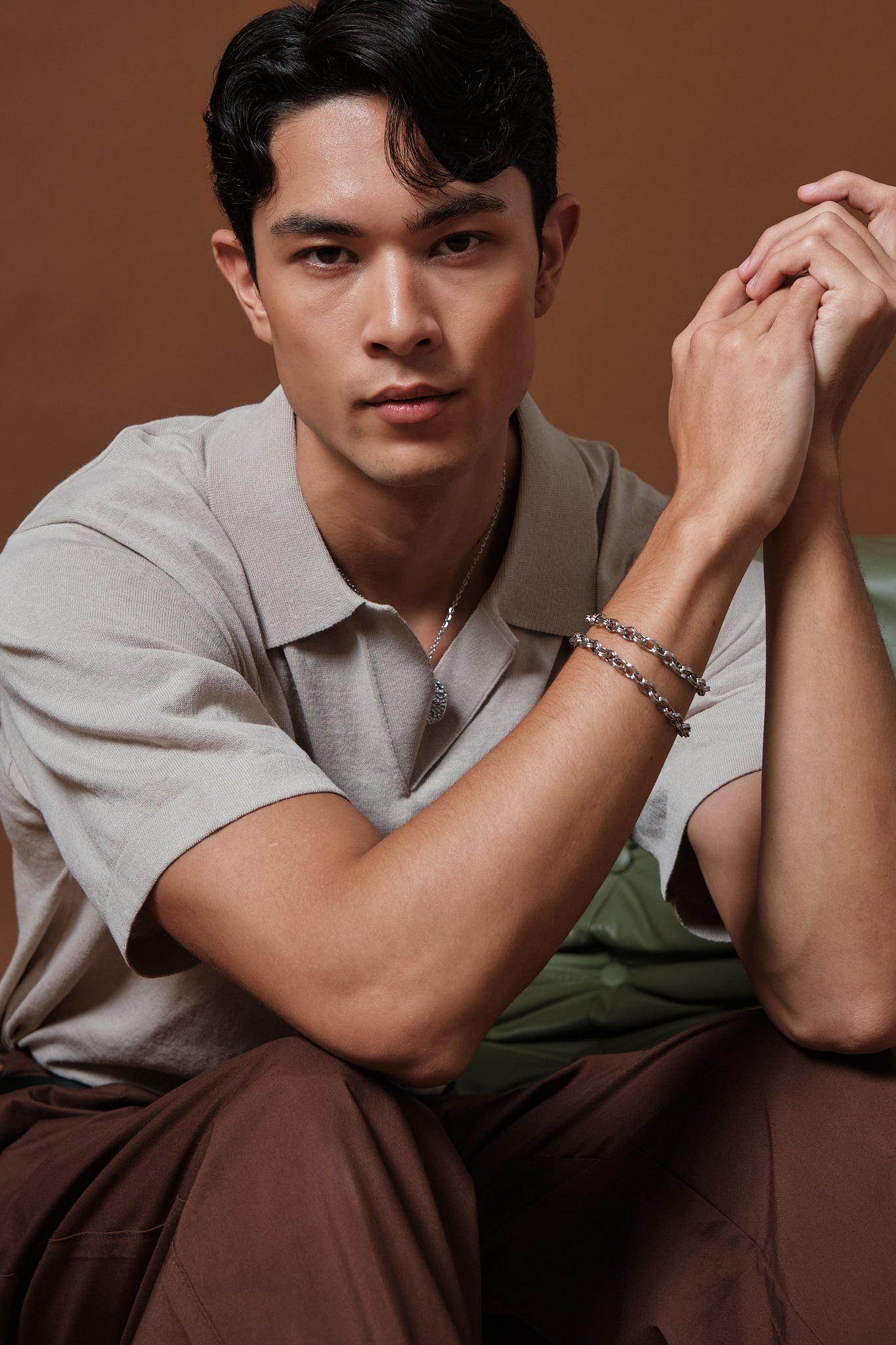 A man wearing a light beige short-sleeve polo shirt and brown trousers, sitting and looking directly at the camera. He is wearing two silver-toned beaded bracelets on his right wrist.
