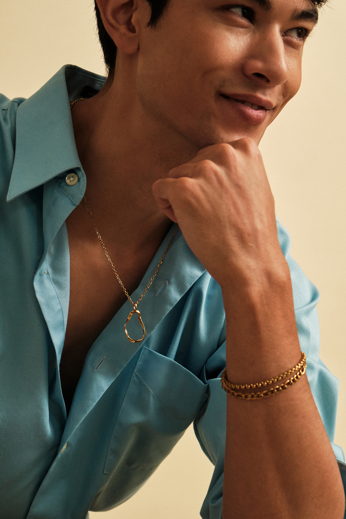 A close-up lifestyle shot of a man wearing a gold infinity loop necklace and a gold chain bracelet, paired with a light blue satin shirt against a warm beige background.