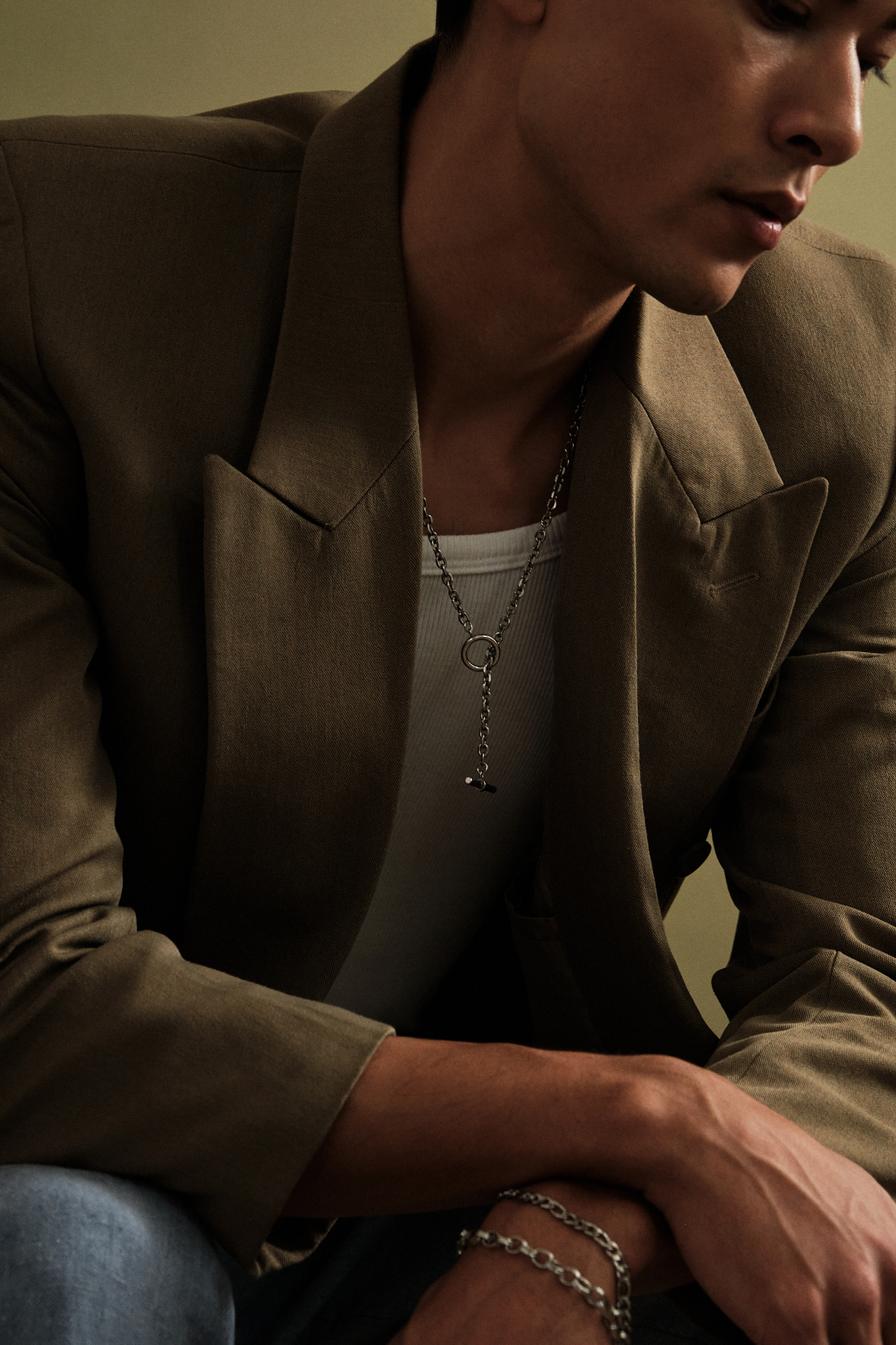 A close-up lifestyle shot of a man wearing a brown blazer, silver chain necklace, and silver bracelets, looking down in a dimly lit setting.