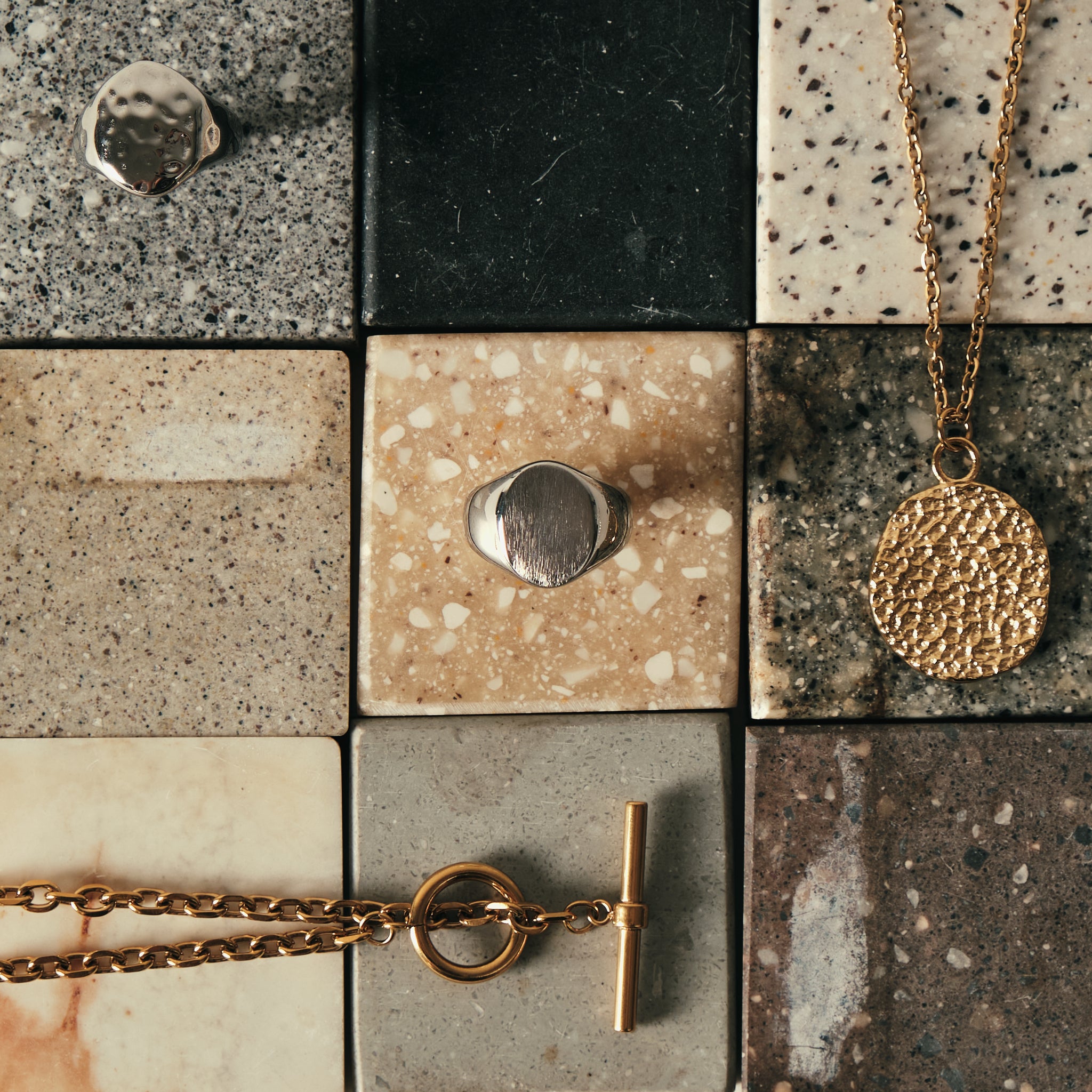 Gold jewelry pieces including a textured round pendant necklace and a toggle ring on a chain, displayed on a grid of speckled and polished stone tiles.