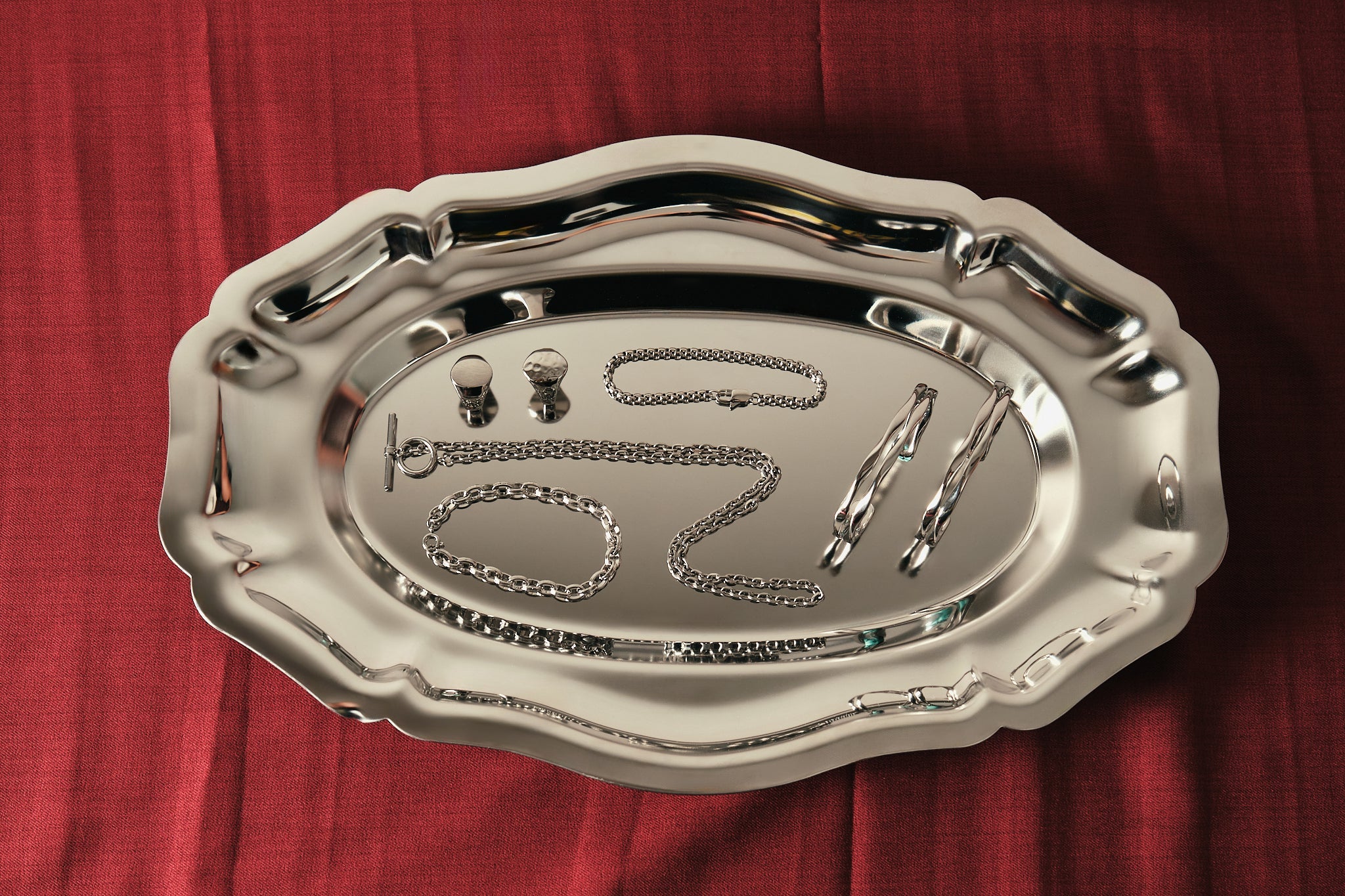 A polished silver metal tray with an ornate scalloped rim, photographed from above on a textured red fabric background. Inside the tray, several small silver jewelry pieces are arranged, including chains and rings.