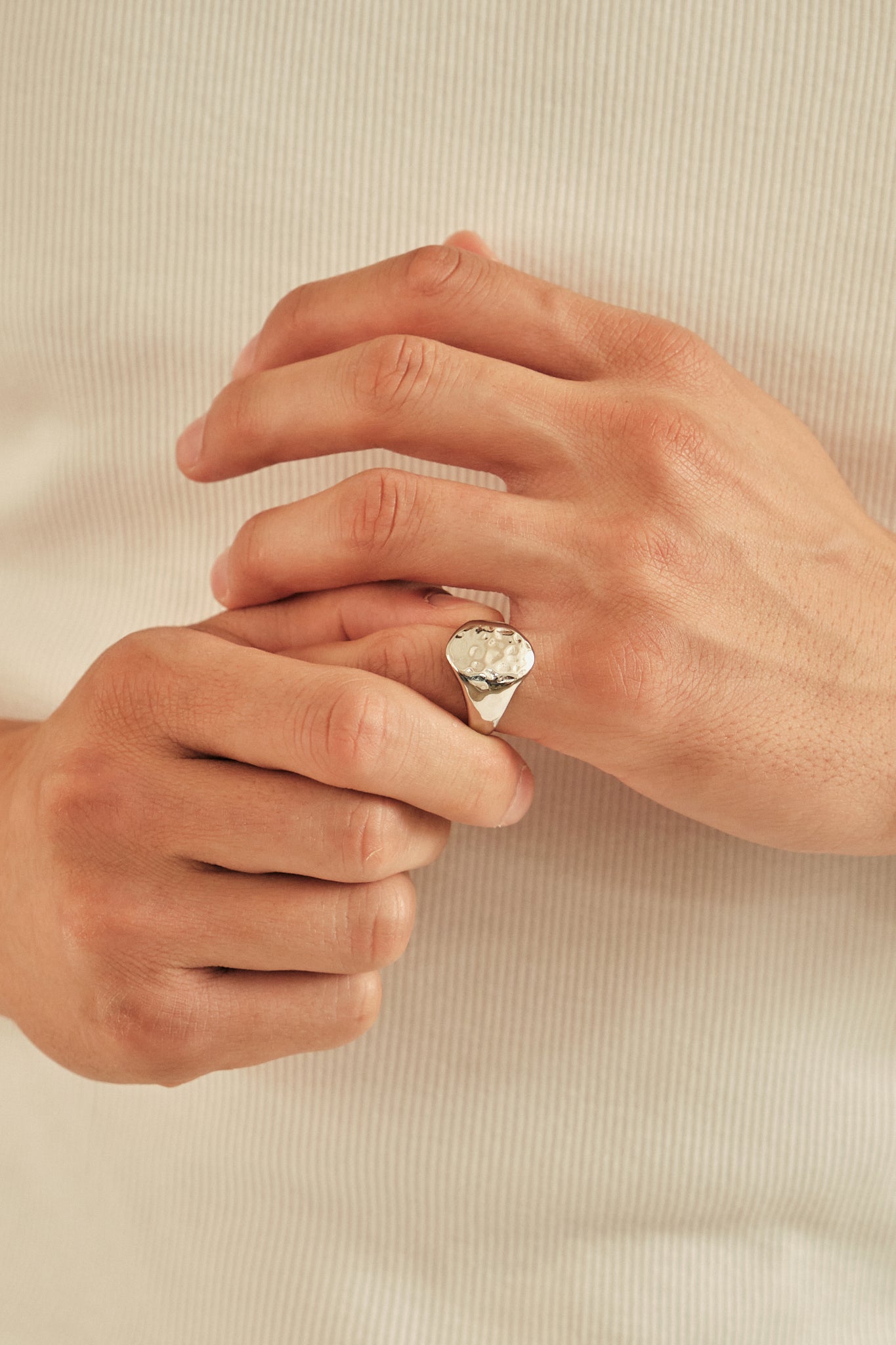 A silver signet ring with a textured, diamond-cut oval face worn on a person's hand, shown against a soft, ribbed cream-colored knit sweater.